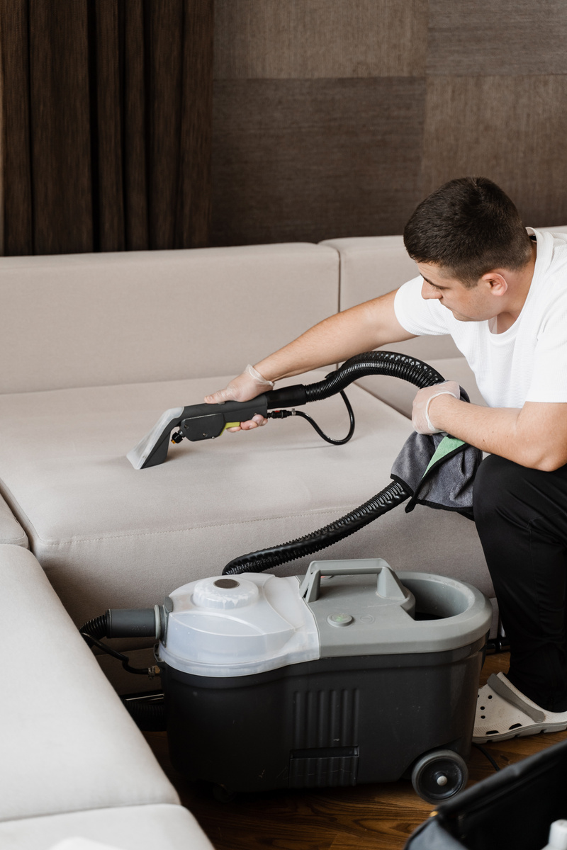 Housekeeper is extracting dirt from upholstered sofa using dry cleaning extractor machine. Cleaning couch with washing vacuum cleaner extraction machine for dry clean upholstered furniture.